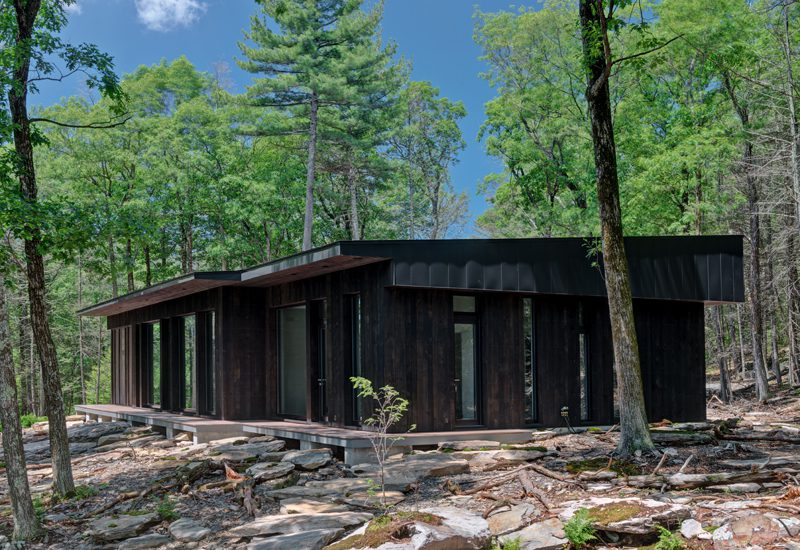 A black cabin sits on top of rocks in the woods.